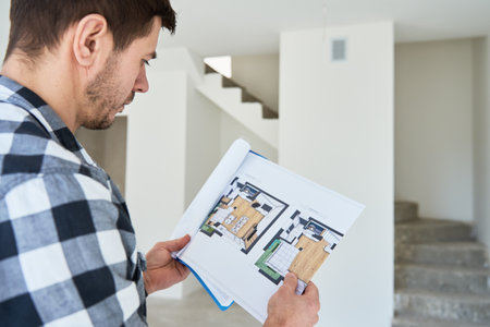 Thoughtful man examines a floor plan while standing in unfinished home. Male home owner analyzing...