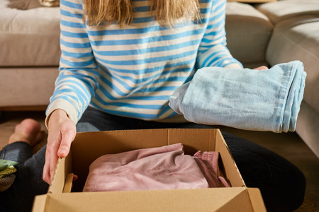 Woman packs cardboard donation box with clothes in living room. Person preparing used clothing for recycling or second hand. Concept of charity, social care and volunteeringの写真素材