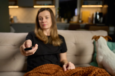 Woman sitting on sofa holding TV remote control. Female watching television at home with pet dog on...