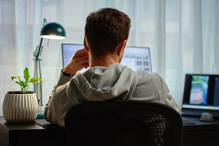 Man in gray hoodie working at computer desk with dual monitors and lamp in background. Freelancer working remotely at home office. Concept of remote work and productivityの写真素材