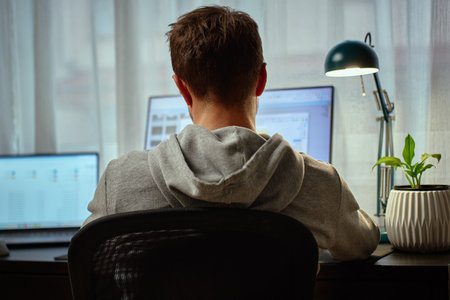 Man in gray hoodie working at computer desk with dual monitors and lamp in background. Freelancer working remotely at home office. Concept of remote work and productivityの写真素材