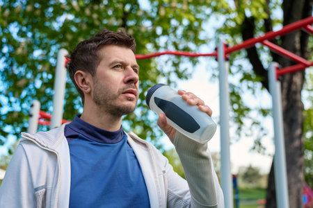 Athletic man drinks water from sports bottle at outdoor gym. Male athlete training in park. Fitness...