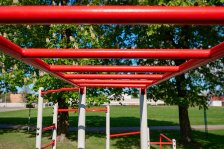Red outdoor monkey bars in park with green trees. Fitness playground horizontal ladder for sport...