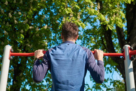 Athletic man performing pull-ups on red horizontal bar in park. Athlete training upper body muscles...