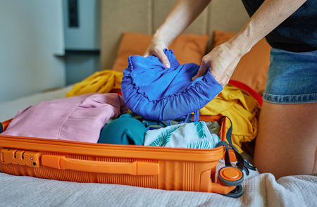 Woman folds colorful clothes into orange suitcase in bedroom. Concept of vacation preparation,...