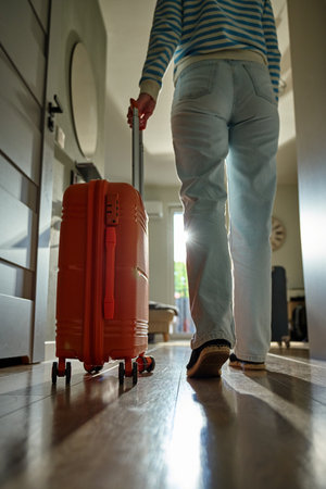 Low-angle view of traveler walking towards door with rolling luggage. Person walks across hallway with orange suitcase. Concept of departure and leaving home for vacationの写真素材