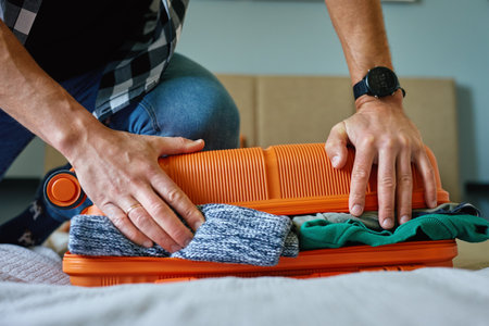 Man presses down on an overfilled orange suitcase while packing clothes. Male traveler has trouble while preparing for travelの写真素材