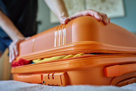 Woman trying to close suitcase at home. Female press down overfilled orange suitcase while packing clothes. Person preparing for departureの写真素材