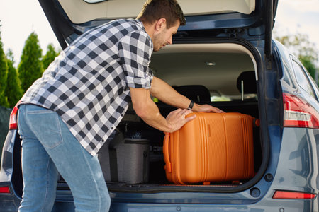 Man loading orange suitcase into car trunk before journey. Packing luggage for travel