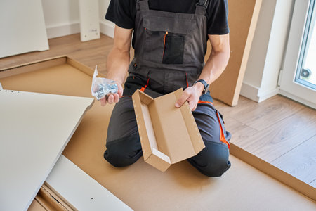 Man in work overalls opens plastic bag with screws and fittings while kneeling beside boxed furniture panels and tools. Furniture assemblyの写真素材