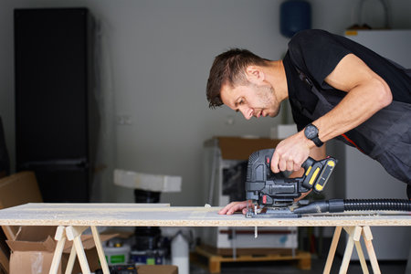 Worker in workwear using handheld cordless electric jigsaw to cut wooden board in garage. Skilled carpenter working in workshop. Man doing DIY home renovationの写真素材