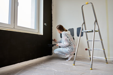 Woman in disposable protective suit painting wall black with brush during home renovation. Concept of gender equality, female empowerment and women doing manual laborの写真素材