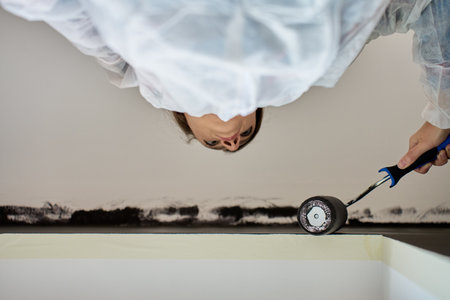 Woman in protective suit painting wall and ceiling with paint roller in black color during interior renovationの写真素材