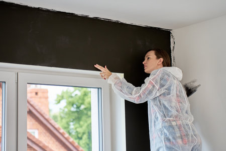 Woman in protective suit inspects freshly painted dark wall in modern apartment. Concept of quality control during DIY renovationの写真素材