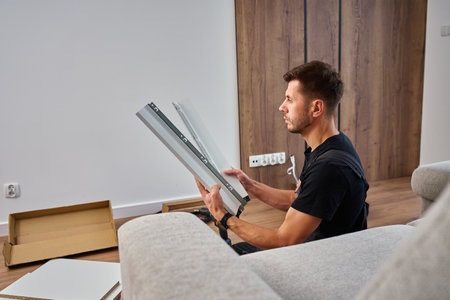 Man in overalls assembles furniture, examining white panel and surrounded by tools. Handyman in work clothes preparing parts pf cabinet to assembly. DIY furniture projectの写真素材