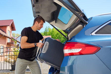 Man loading suitcase into car trunk on sunny day. Concept of preparing for summer travel, vacation or road trip with luggageの写真素材