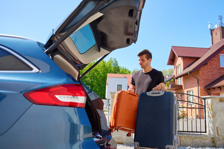 Man holding orange and blue suitcases near parked car in front of suburban brick house. Loading luggage in vehicle. Concept of moving day, relocation or travel preparationの写真素材