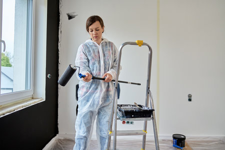 Woman wearing protective coveralls holds black paint roller near painted wall samples and ladder in room under renovation. Painter preparing to paint wall. Home improvement and interior update conceptの写真素材