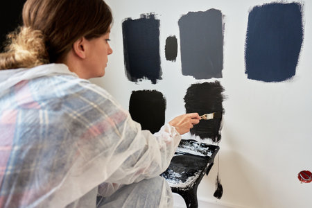 Woman in protective wear applies different dark paint swatches on white wall using brush. Female painter testing black and dark gray paint samples during interior renovation project in modern homeの写真素材
