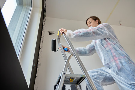 Woman in protective wear climb ladder with paint roller in hand near window, preparing to paint black accent wall. Home renovation in modern apartment interiorの写真素材