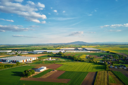 Aerial view of industrial warehouse complex near green agricultural fields and small village. Modern...