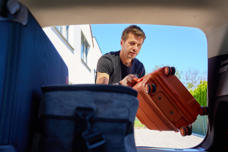 Close up of man loading orange suitcase into open car trunk outside residential house. Inside view of male traveler placing luggage in vehicle before road trip. Preparation before travel or departureの写真素材