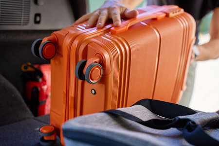 Close up of hand placing orange suitcase into car trunk, preparing for travel or vacation. Inside view of man loading luggage in vehicleの写真素材