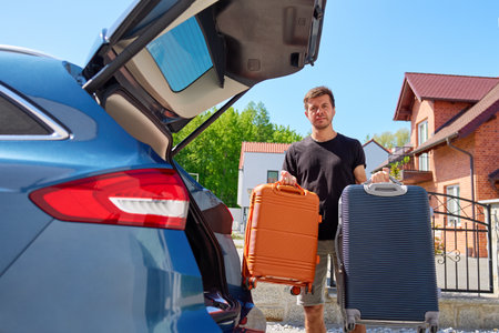 Man holding two suitcases near car with open trunk parked in front of brick house. Male driver preparing for loading luggage into vehicle. Concept of relocation or moving to new homeの写真素材