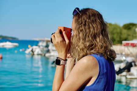 Woman photographing with instant camera near marina with boats in background. Female tourist taking photo by sea water with retro camera Summer vacation and travel conceptの写真素材