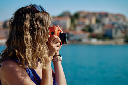 Woman holding orange instant camera, taking photo near sea with blurred town in background. Female tourist using retro camera during summer travel. Summer tourism and holiday conceptの写真素材