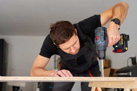 Worker using power drill on wooden board supported by sawhorses in home garage workshop. Man working in workshop, drilling hole in chipboard for furniture assemblyの写真素材
