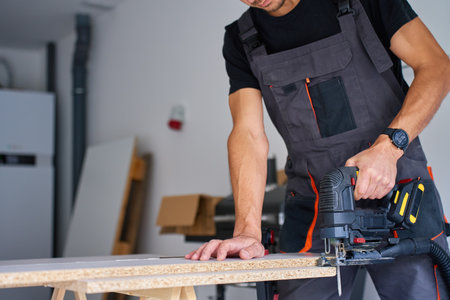Carpenter cuts wooden board with electric jigsaw in home workshop. Man in workwear working with wood on workbench in garage. Woodworking and carpentry conceptの写真素材