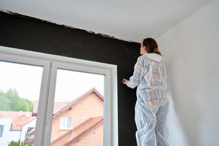 Woman in protective suit inspects freshly painted dark wall in modern apartment. Concept of quality control during DIY renovationの写真素材