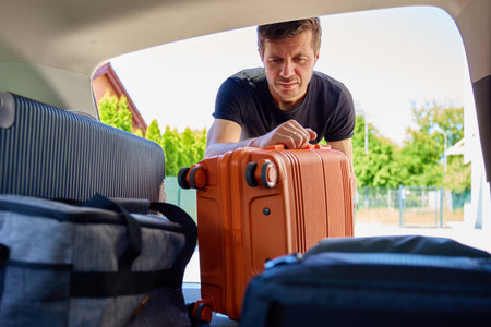 Man placing orange rolling suitcase into open vehicle trunk, preparing for journey or vacation on sunny day. Inside view of traveler loading luggage in car. Travel or weekend road trip conceptの写真素材