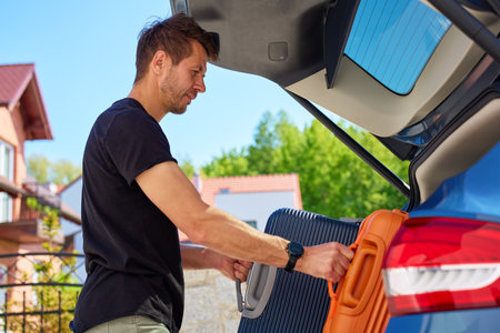 Man loading orange suitcase into car trunk on sunny day. Concept of preparing for summer travel, vacation or road trip with luggageの写真素材