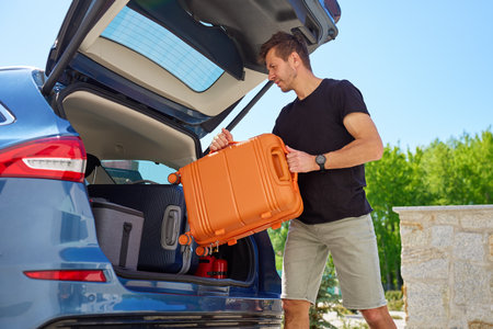 Man loading orange suitcase into car trunk on sunny day. Concept of preparing for summer travel, vacation or road trip with luggageの写真素材