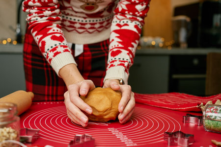 Woman in cozy red sweater prepares gingerbread dough for baking holiday cookies in kitchen. Close-up of hands kneading dough on red mat surrounded by cookie cutters and Christmas decorationsの写真素材