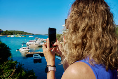 Rear view of woman in blue dress taking picture with smartphone near sea on sunny day. Female tourist photographing seascape with phone. Summer vacation, travel and technology conceptの写真素材