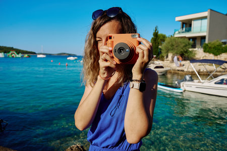 Woman photographing with instant camera near marina with boats in background. Female tourist taking photo by sea water with retro camera Summer vacation and travel conceptの写真素材