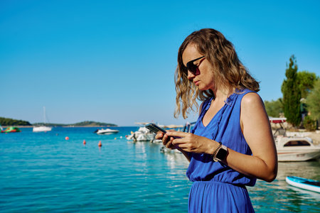 Woman in sunglasses and blue dress using smartphone by sea on sunny day. Female tourist texting by waterfront. Concept of digital lifestyle during travelの写真素材