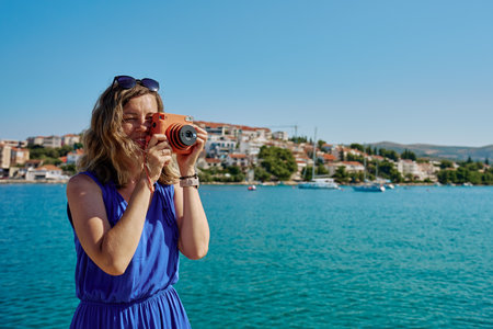 Woman standing near sea holding instant camera and taking photo. Female tourist at coastal resort with blurred town in background. Travel and summer vacation conceptの写真素材