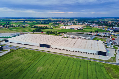 Large modern warehouse with loading docks and parked trucks surrounded by green fields. Industrial logistics hub. Transport infrastructure for distribution, shipping and storage in supply chain systemの写真素材