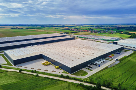 Large modern warehouse with loading docks and parked trucks surrounded by green fields. Industrial logistics hub. Transport infrastructure for distribution, shipping and storage in supply chain systemの写真素材