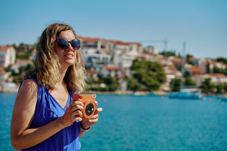 Woman holding orange instant camera, taking photo near sea with blurred town in background. Female tourist using retro camera during summer travel. Summer tourism and holiday conceptの写真素材