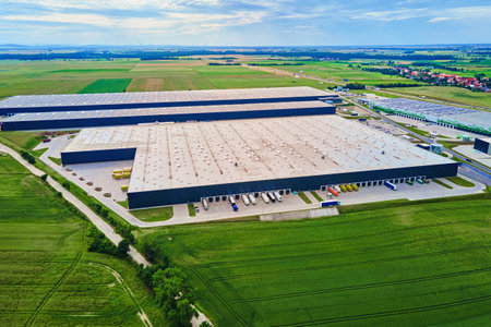 Large modern warehouse with loading docks and parked trucks surrounded by green fields. Industrial...