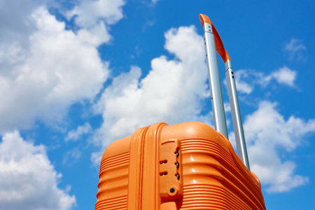 Bright orange suitcase with extended handle stands against blue sky with white clouds. Concept of travel, vacation and summer adventureの写真素材