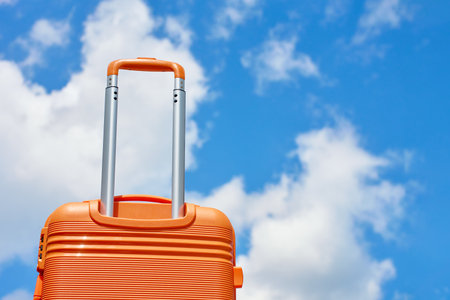 Bright orange suitcase with extended handle stands against blue sky with white clouds. Concept of travel, vacation and summer adventureの写真素材
