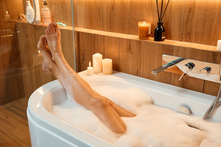Female legs in bathtub filled with foam and warm lighting. Woman relaxing in bathroom with candles. Concept of wellness, relaxation, and self careの写真素材