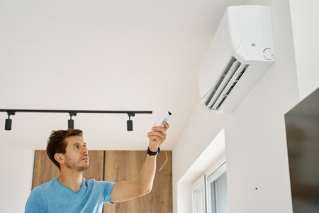 Man using remote control to adjust temperature on wall-mounted air conditioner in modern home interior. Concept of home cooling and indoor climate control during hot summer weatherの写真素材