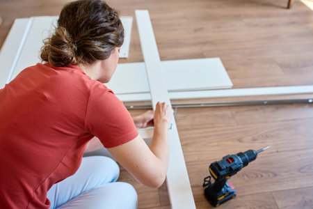 Woman sits on wooden floor and assembling furniture in new home, fastens parts of white bed frame. DIY home renovation and home organization after moving conceptの写真素材